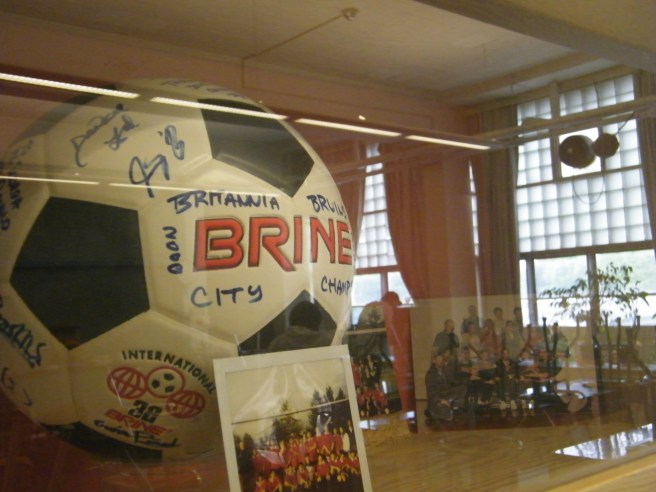 A glassed-in shelf, holding a signed soccer ball with photos and memorabilia. The reflection of a classroom can be seen in the glass, especially the large, paned windows. A glassed-in shelf, holding a signed soccer ball with photos and memorabilia. The reflection of a classroom can be seen in the glass, especially the large, paned windows.