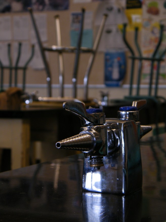 A table in a science classroom, with a gas nozzle in the foreground, stools stacked on desks in the background. A table in a science classroom, with a gas nozzle in the foreground, stools stacked on desks in the background.