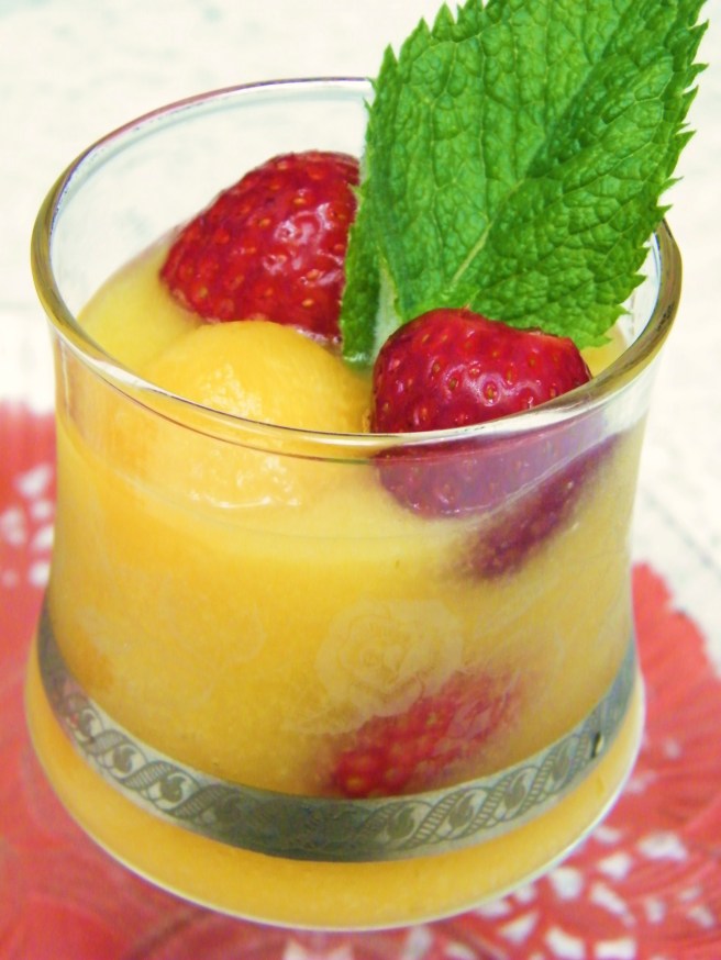 An etched wine glass full of melon soup, with melon balls and strawberries and a sprig of mint. Everything sits on a tray atop a red doily on a white lace tablecloth. An etched wine glass full of melon soup, with melon balls and strawberries and a sprig of mint. Everything sits on a tray atop a red doily on a white lace tablecloth.