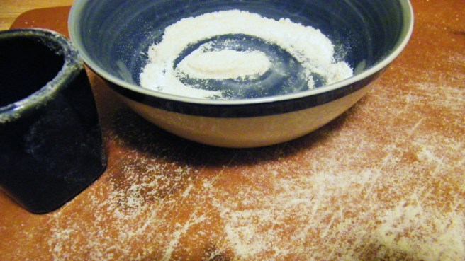 A floured board, a blue-and-white bowl with flour in it, and a Japanese tea cup subbing as a biscuit-cutter