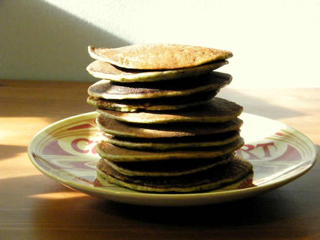 A stack of Swiss chard pancakes on a yellow and red plate that says Camembert, atop a wooden table with light and shadows cutting across the frame.